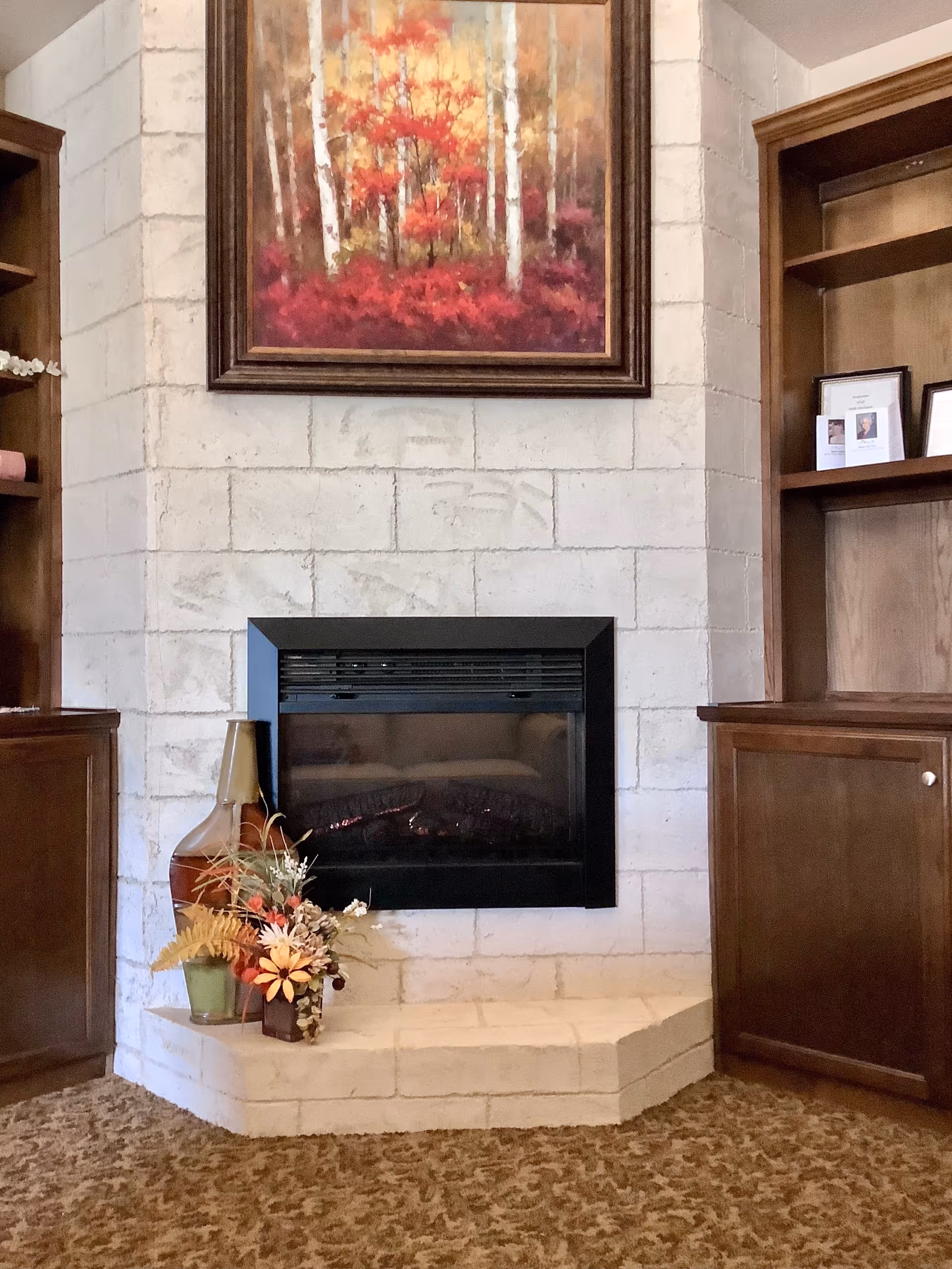 A white brick fireplace with a black electric insert topped by an autumn forest painting, flanked by wooden built-in shelves and decorative vases and flowers on the hearth.