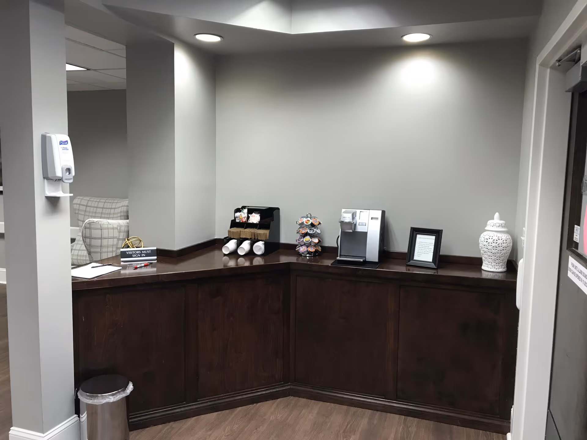 Reception or check-in counter area with a dark wood countertop and cabinets below. On the counter are a coffee machine, a carousel with coffee pods, a small framed sign, a decorative white ceramic jar, and a sign that reads 'VISITORS MUST SIGN IN'. A hand sanitizer dispenser is mounted on the wall to the left. In the background, there is a glimpse of a seating area with a plaid armchair.