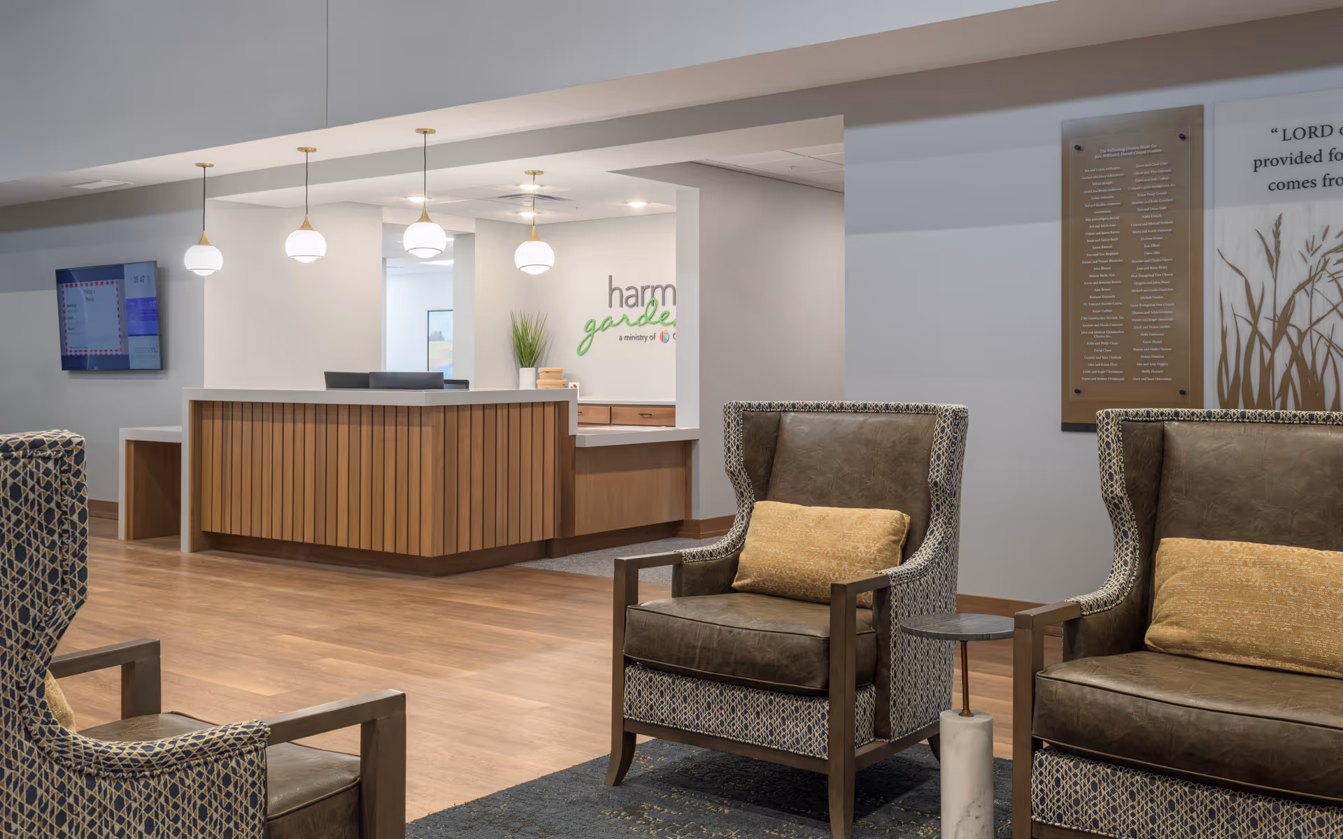 A modern senior living facility reception area with a wooden front desk, three hanging globe pendant lights, and a wall-mounted TV. Two upholstered armchairs with patterned fabric and yellow cushions are positioned in the foreground on a rug. The wall behind the desk features the Harmony Gardens logo and some framed artwork.