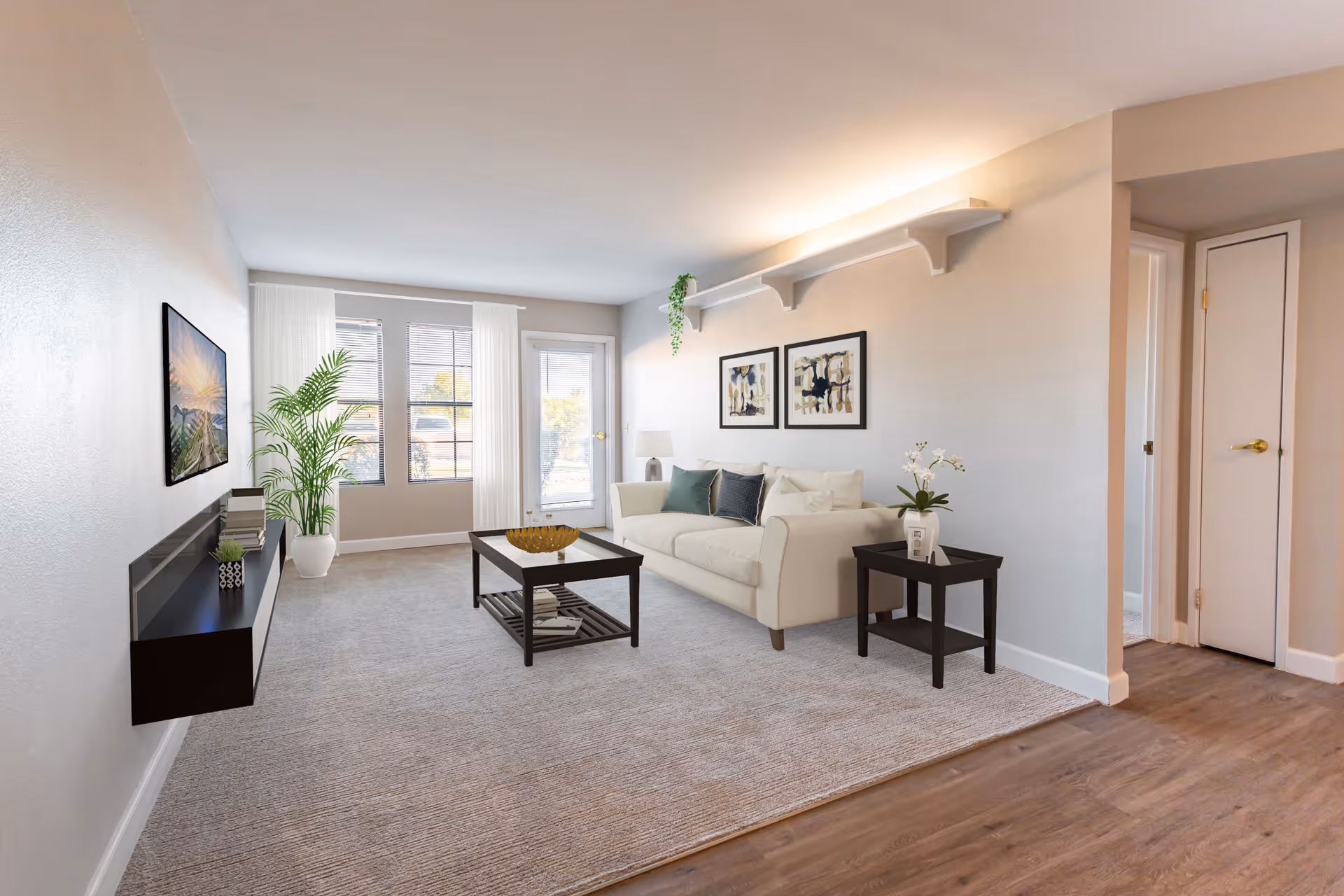 A bright and modern living room with a white sofa adorned with cushions, a dark wooden coffee table with a decorative bowl, and a matching side table with a potted plant. The room features a large window and glass door letting in natural light, a wall-mounted flat-screen TV, a floating shelf with books and plants, and two framed abstract paintings on the wall.