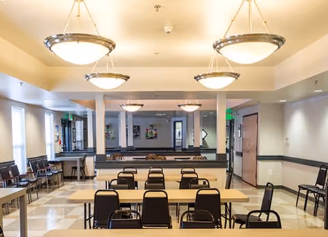A well-lit dining area with multiple rectangular tables and black chairs arranged in rows. The room features large hanging ceiling lights, windows on the left side, and light-colored walls with a dark chair rail. There are doors and additional seating visible in the background.