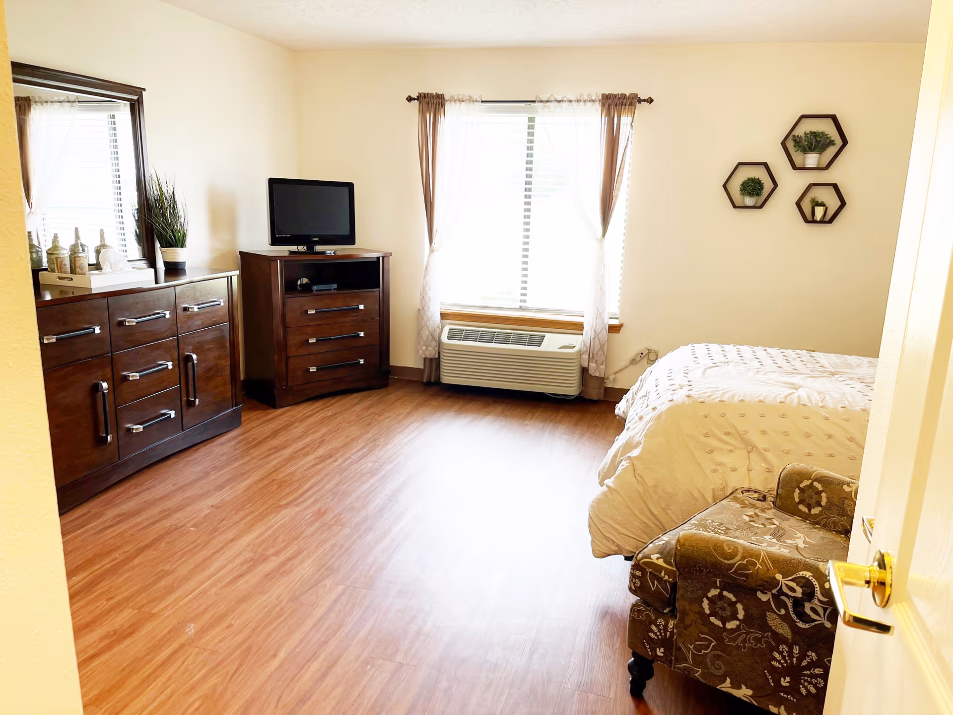 A bright bedroom with wooden flooring featuring a bed with white bedding, a patterned armchair, a wooden dresser with a mirror, a small wooden chest with a TV on top, and a window with sheer curtains. Three hexagonal wall shelves with small green plants are mounted on the wall.