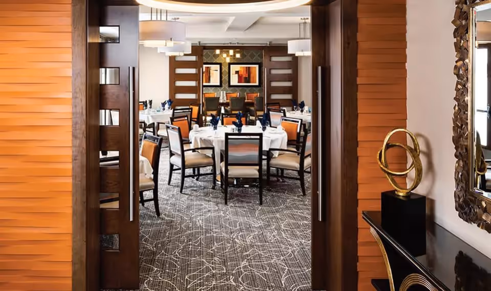View through open double wooden doors into a dining room with round tables covered in white tablecloths, set with blue napkins and chairs with orange and beige cushions. The room features modern light fixtures, patterned carpet, and framed artwork on the far wall. To the right, there is a decorative gold sculpture on a black pedestal and a large ornate mirror on the wall.