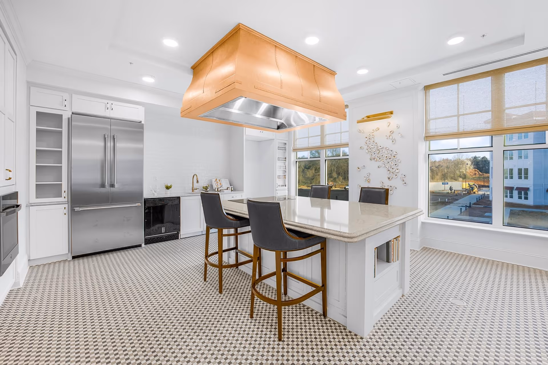 Bright and modern kitchen with a large island in the center featuring a light-colored countertop and three high chairs. The kitchen has white cabinetry, a stainless steel refrigerator, built-in oven, and a copper range hood above the island. Large windows with beige roller shades allow natural light to fill the space, and the floor is covered with a patterned tile.