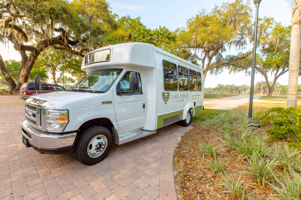 A white shuttle bus parked on a paved driveway surrounded by greenery and trees. The bus has the logo and name 'Lake Gibson Village' on its side, indicating it is part of a senior living community. The background shows a clear sky and a landscaped area with plants and trees.
