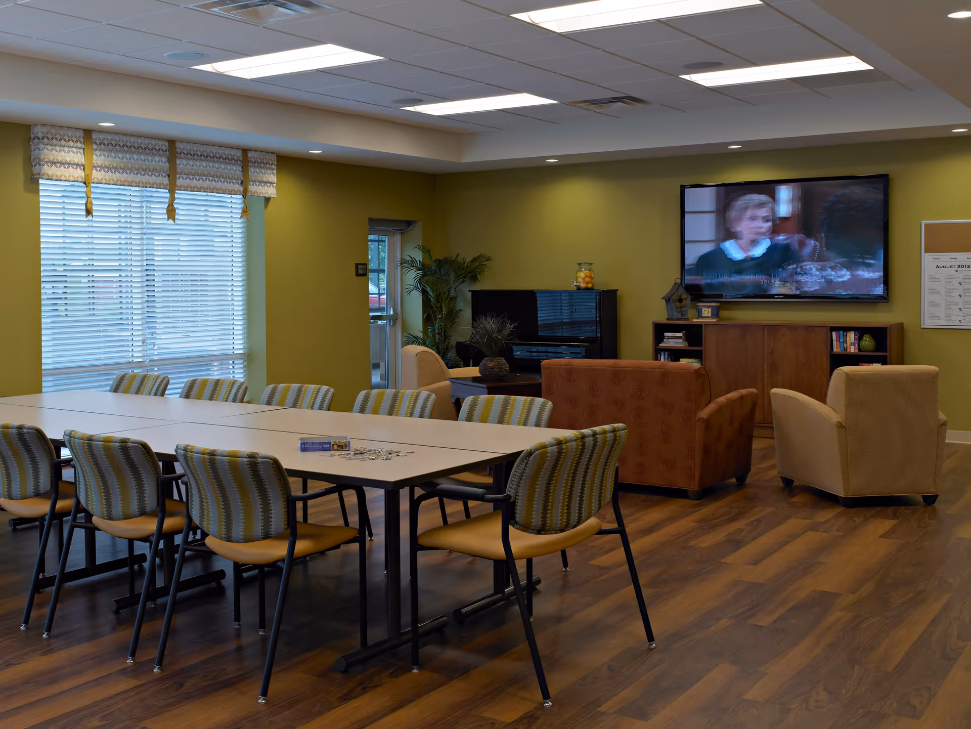 A spacious room with a long table surrounded by chairs with patterned upholstery. In the background, there is a seating area with armchairs facing a wall-mounted television. The room has green walls, large windows with blinds and valances, and wood flooring. A piano and a cabinet with books and decorative items are also visible.