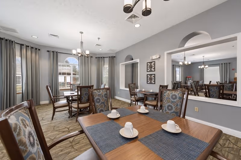 A dining room with multiple wooden tables and chairs featuring floral patterned upholstery. Each table is set with white cups and saucers on blue placemats. The room has gray walls, large windows with gray curtains, a carpeted floor, and modern light fixtures hanging from the ceiling. A large mirror on one wall reflects the room.