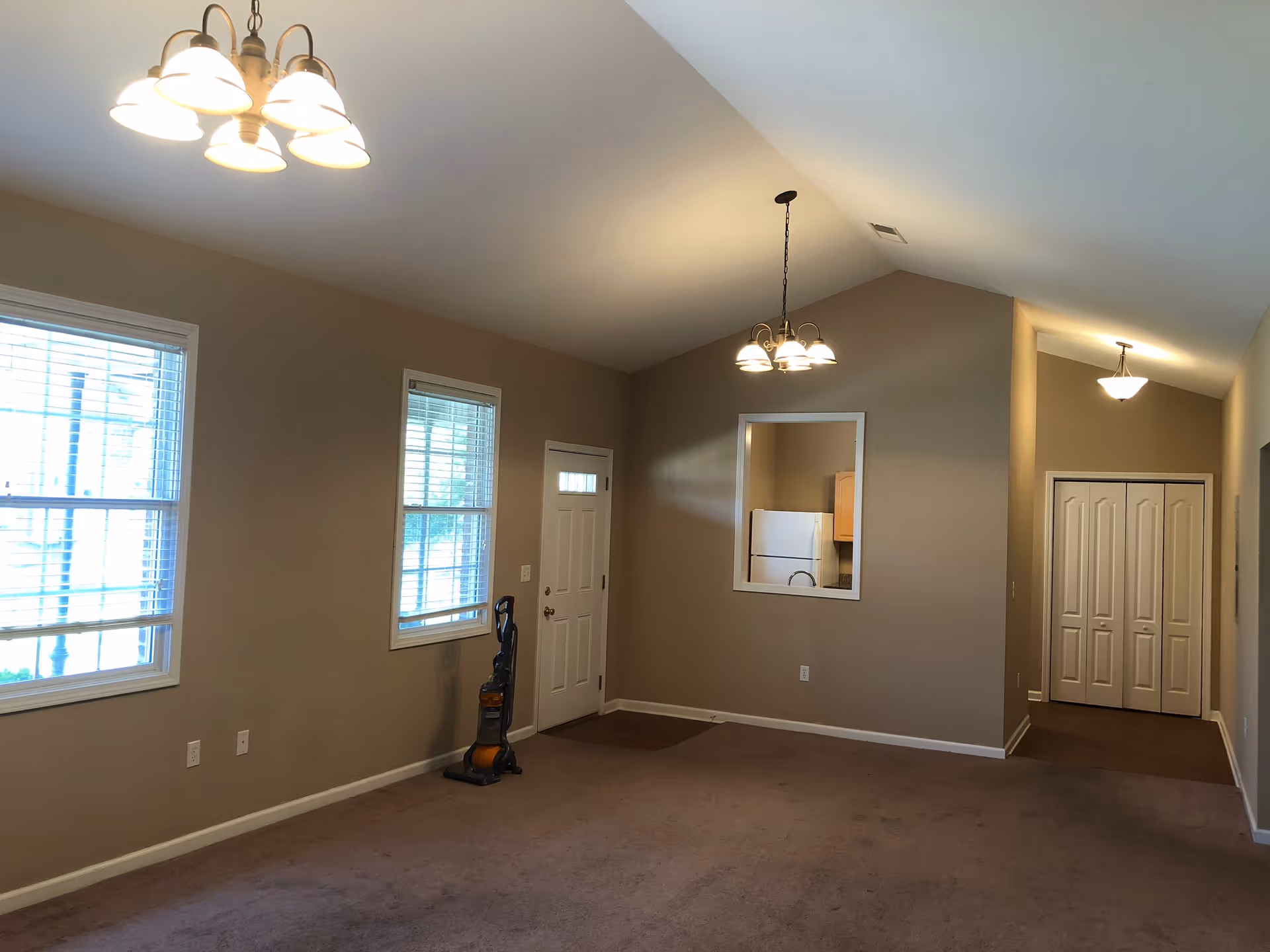 An empty living room area with beige walls and carpeted floor. There are two windows with white blinds on the left wall, a white front door between the windows, and a vacuum cleaner standing near the door. The ceiling is vaulted with three light fixtures hanging down. A pass-through window reveals a kitchen area with a refrigerator. At the far end, there is a hallway with a double-door closet.