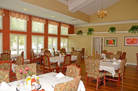 A bright dining room with multiple tables covered in white tablecloths and set with condiments and napkins. The room features large windows with valances, floral upholstered chairs, hardwood floors, hanging chandeliers, and framed floral artwork on the walls. Green plants are placed on a high ledge near the ceiling.