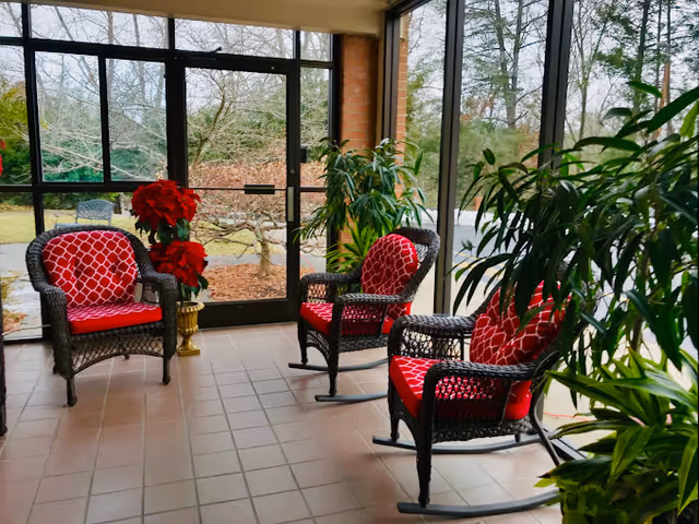 A sunroom or enclosed porch area with three black wicker rocking chairs featuring red cushions with a white geometric pattern. There are several green potted plants around the chairs, and a large window and glass door provide a view of the outdoor garden area with trees and shrubs.