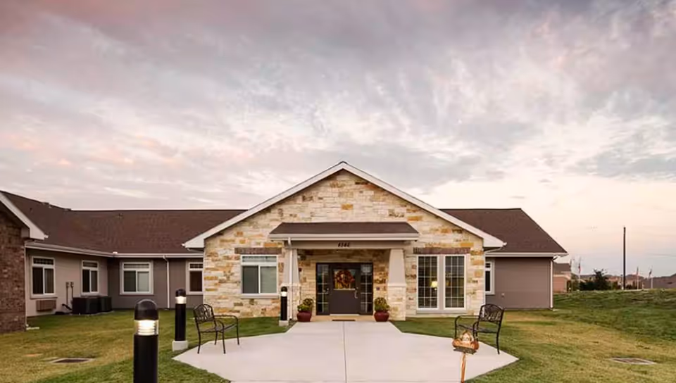 Exterior view of a single-story building with a stone facade and a brown roof, featuring a central entrance with a small covered porch. There are benches and lamp posts along the concrete pathway leading to the entrance, with grass on either side and a partly cloudy sky at dusk.
