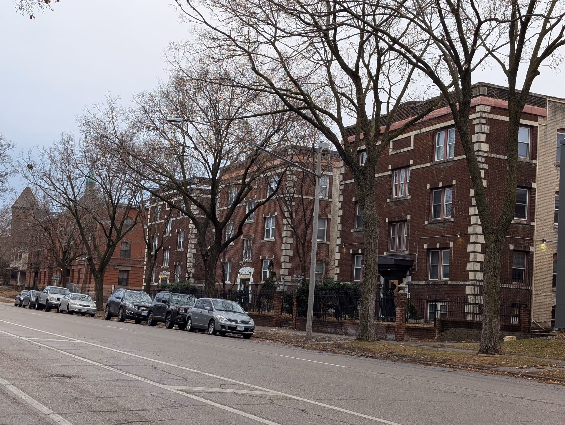 Street view of a residential area with several parked cars along the curb and multiple brick apartment buildings with leafless trees in front during a cloudy day.