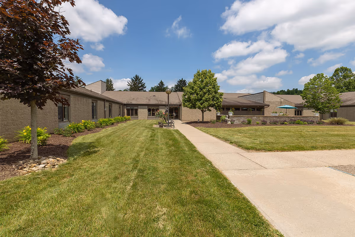 Exterior view of Adams Lane Healthcare & Rehabilitation building with a well-maintained lawn, trees, and a paved walkway leading to the entrance under a partly cloudy sky.