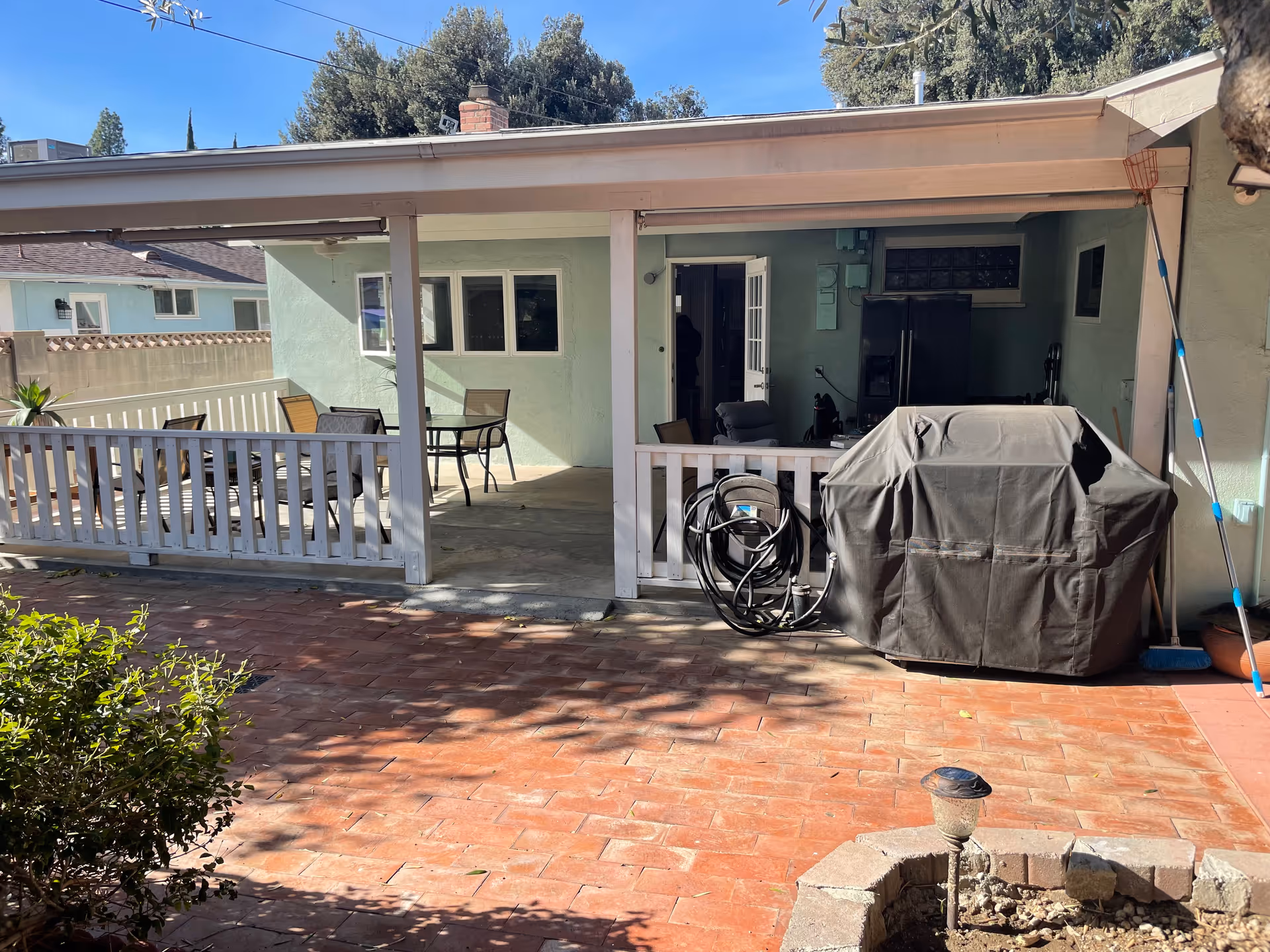 Outdoor patio area with a covered seating space featuring a table and chairs. There is a covered grill on the right side, a coiled hose hanging on the railing, and a small garden bed with a solar light in the foreground. The building exterior is light green with white trim and a clear blue sky above.