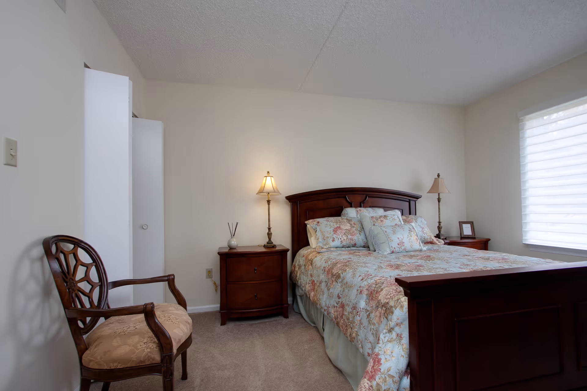 A bedroom with a wooden bed frame and floral bedding. There are two matching wooden nightstands on either side of the bed, each with a lamp. A wooden chair with a cushioned seat is positioned near the left wall. A window with a white blind is on the right side of the room, allowing natural light to enter.