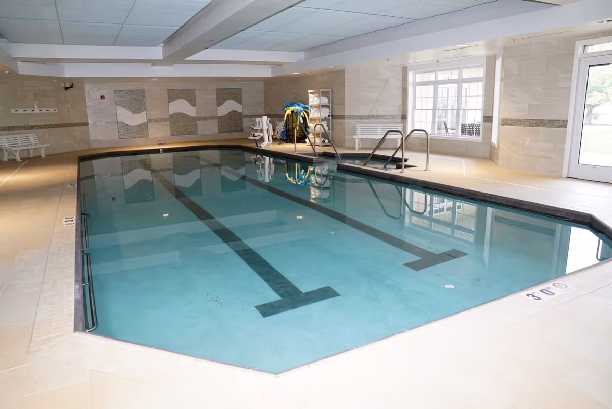 Indoor swimming pool with clear blue water, surrounded by beige tiled flooring and walls. There are benches along the walls, pool safety equipment, and large windows letting in natural light.