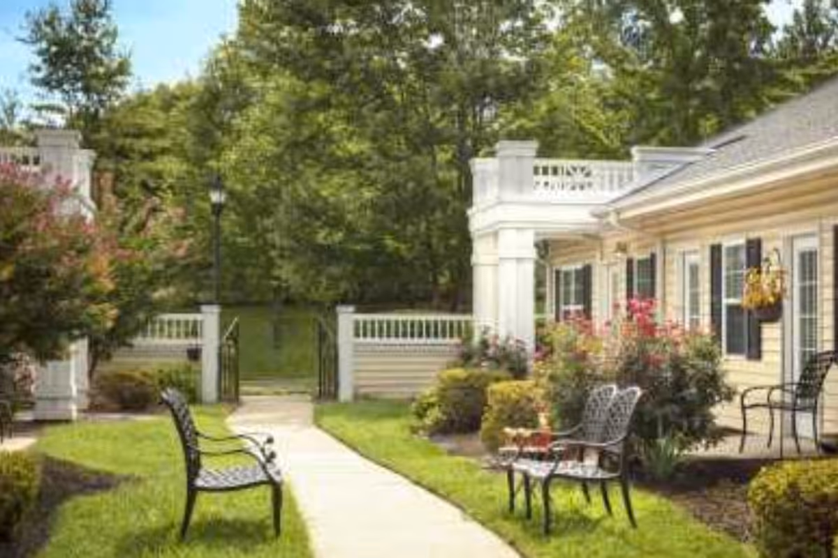 Outdoor garden area at Harmony House Memory Care with a paved walkway, black metal benches, green grass, bushes, flowering plants, and a white building with black shutters and hanging flower pots in the background.