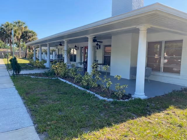 A long covered porch with white columns and ceiling fans attached to a white brick building. The porch has several chairs and small tables, with a garden bed containing small shrubs and plants running alongside it. Palm trees and a sidewalk are visible on the left side of the image.