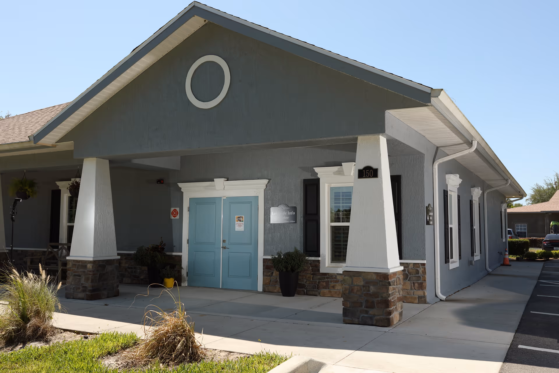 Exterior view of a single-story building with a covered entrance supported by two white pillars with stone bases. The building is painted gray with white trim and has a pair of light blue double doors at the entrance. There is a small sign next to the door and a black plaque with the number 150 on one of the pillars. The surrounding area includes a sidewalk, some plants, and a parking lot.