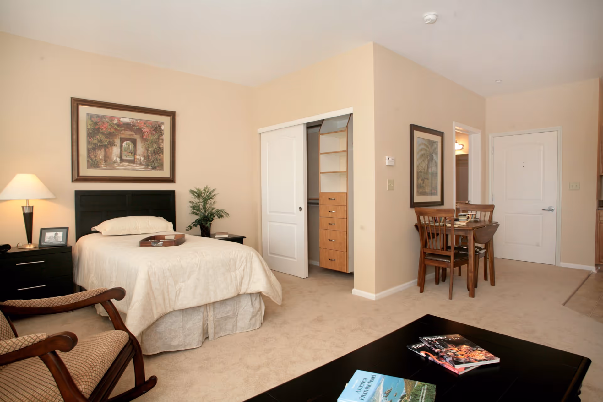 A cozy senior living apartment featuring a single bed with beige bedding, a bedside table with a lamp and framed photo, a wooden armchair, a small dining table with two chairs, and an open closet with built-in shelves. The walls are painted light beige and decorated with framed artwork. A coffee table with magazines is in the foreground.
