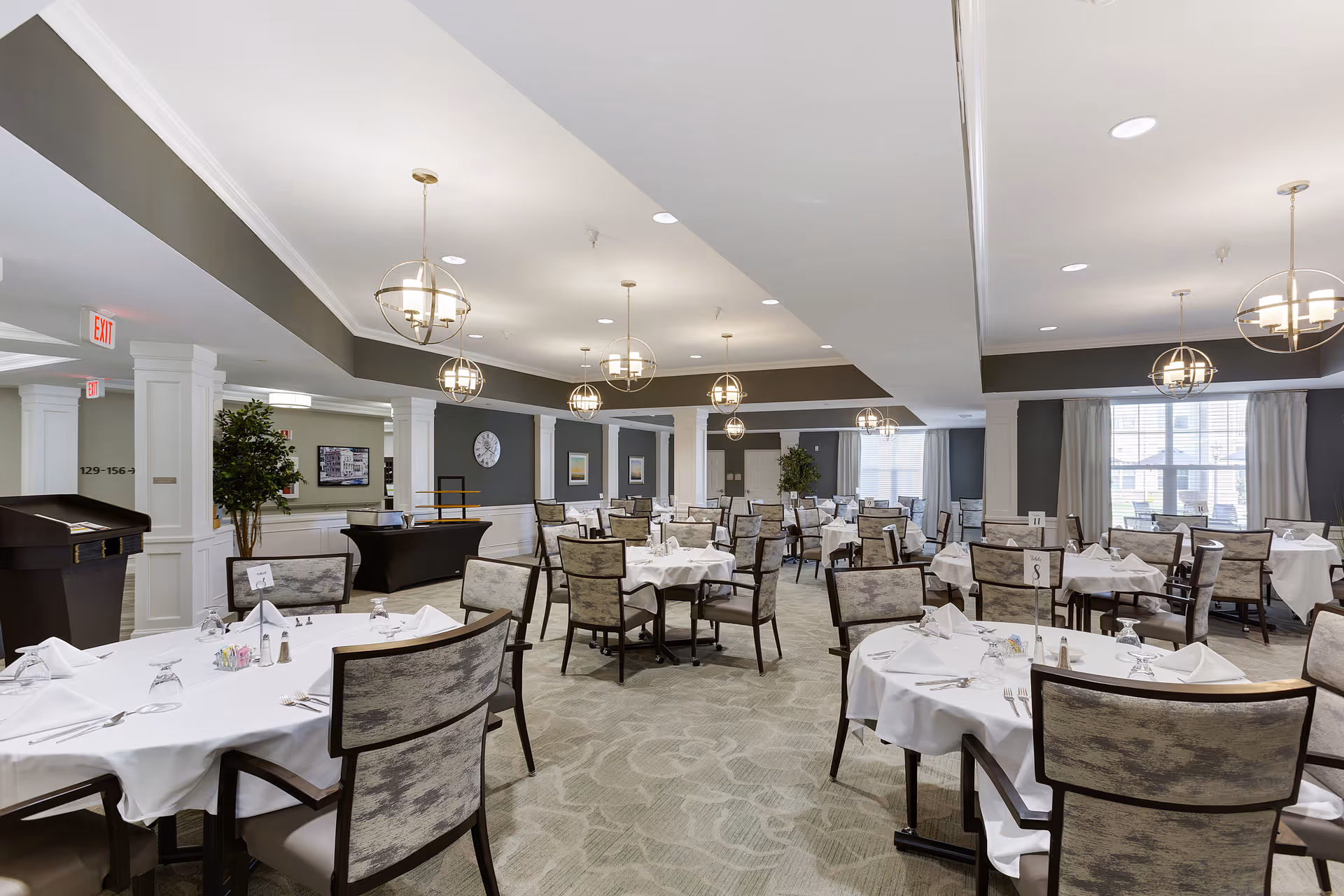 A spacious dining room with multiple round tables covered with white tablecloths and set with napkins, glasses, and silverware. The room features modern hanging light fixtures, large windows with curtains, and neutral-colored walls with decorative columns and framed artwork.