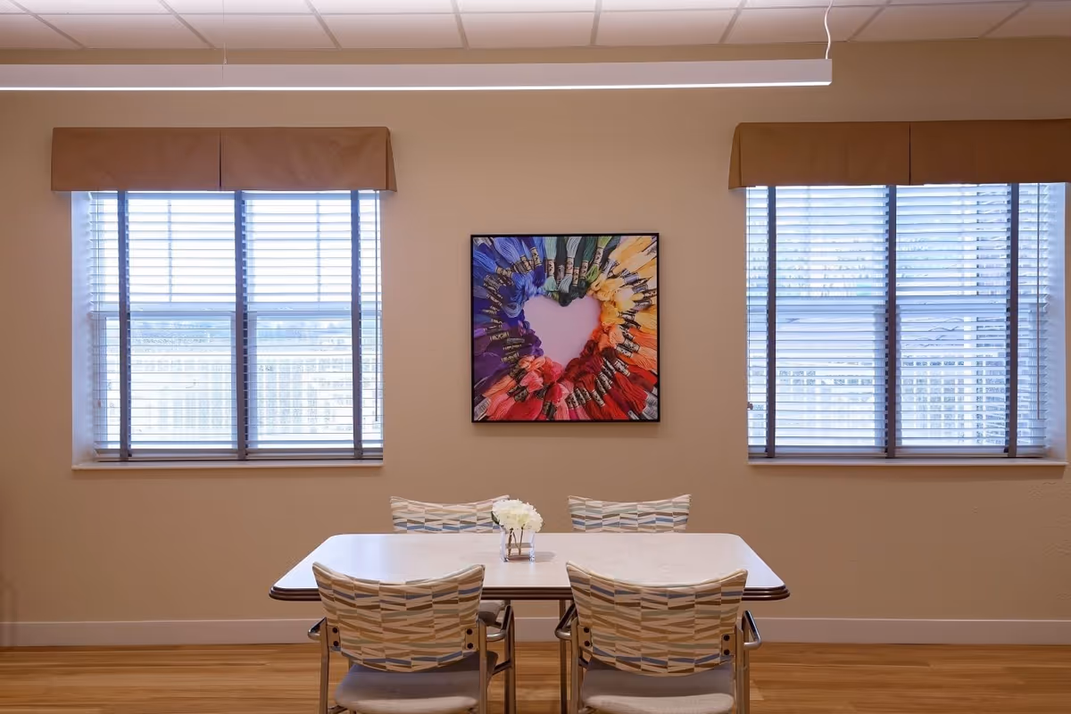 A small dining area with a rectangular table and four patterned chairs. The table has a small vase with white flowers. Two large windows with blinds and brown valances flank a colorful framed artwork on the wall depicting embroidery floss arranged in a heart shape.