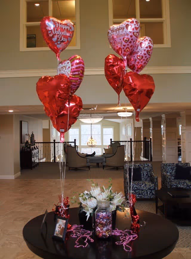 Lobby interior decorated for Valentine's Day with heart-shaped balloons above a table holding a floral centerpiece and candies.