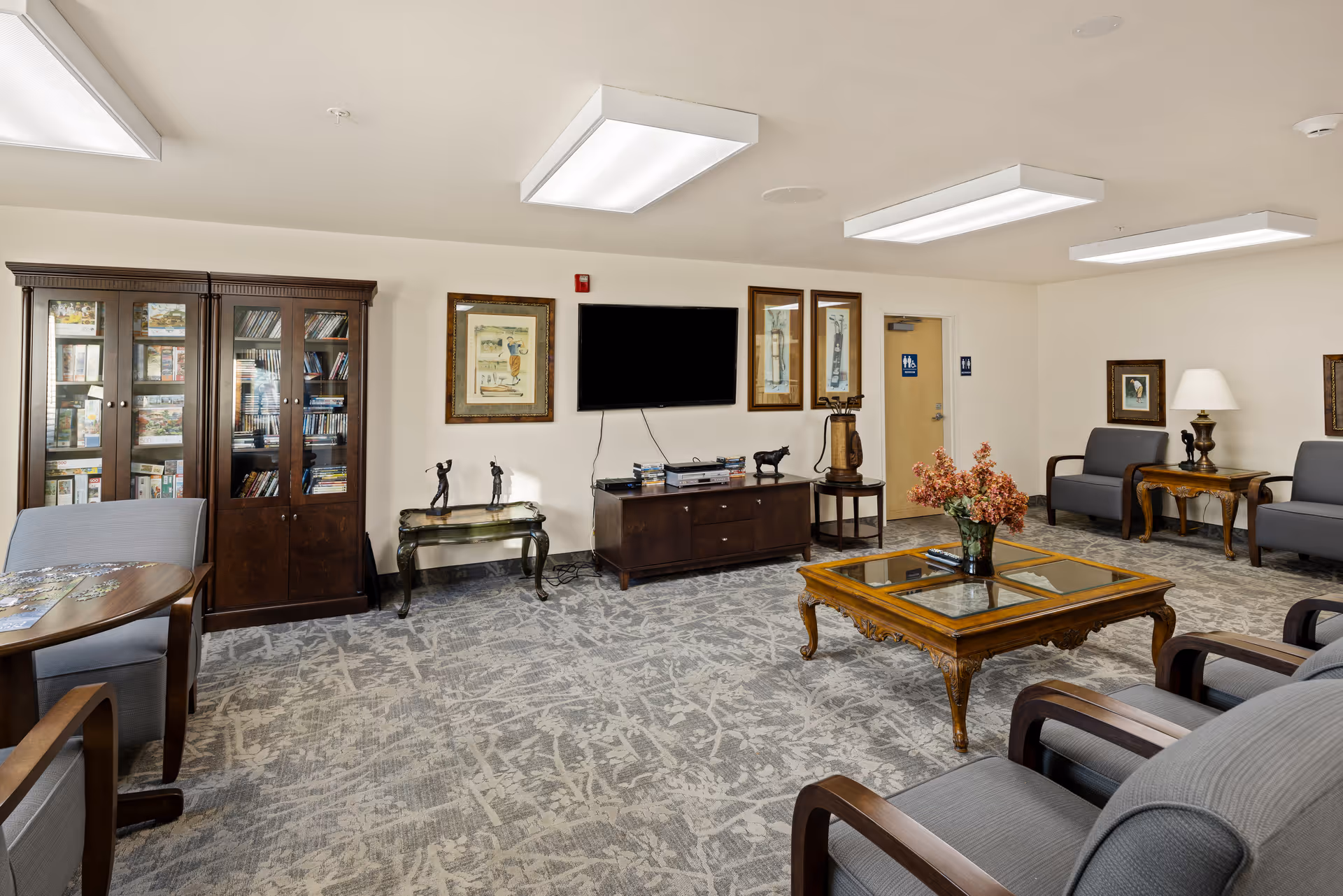 A senior living community lounge with gray upholstered chairs and wooden armrests arranged around a wooden coffee table with a glass top and a vase of flowers. The room features a patterned carpet, a wooden cabinet with glass doors filled with books and games, a wall-mounted flat-screen TV, framed artwork, and a side table with a lamp. There is a door with restroom signs in the background.