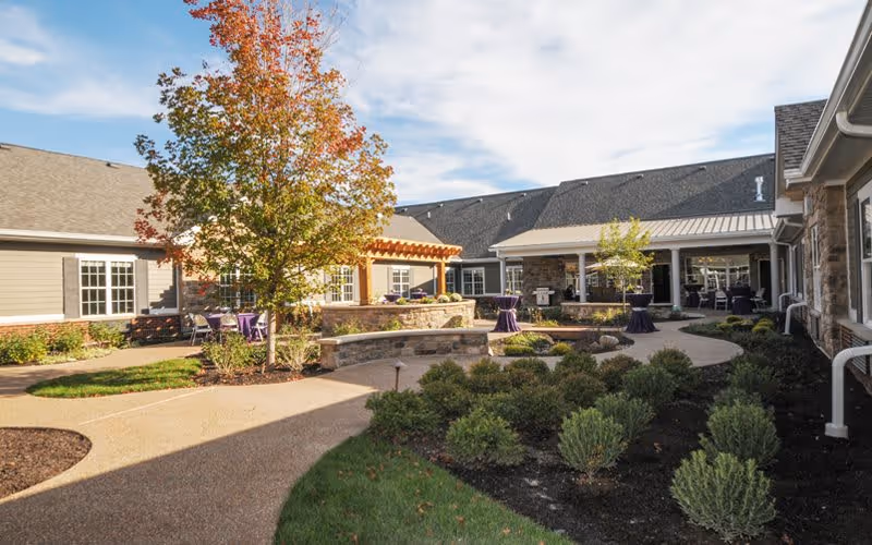 Outdoor courtyard area of a senior living facility with a paved walkway, landscaped bushes, a tree with autumn leaves, and seating areas under a pergola and covered patio.