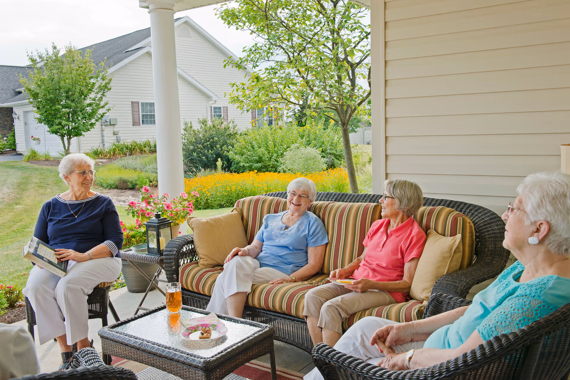 Four elderly women sitting and chatting on a covered outdoor patio with wicker furniture and a garden in the background.