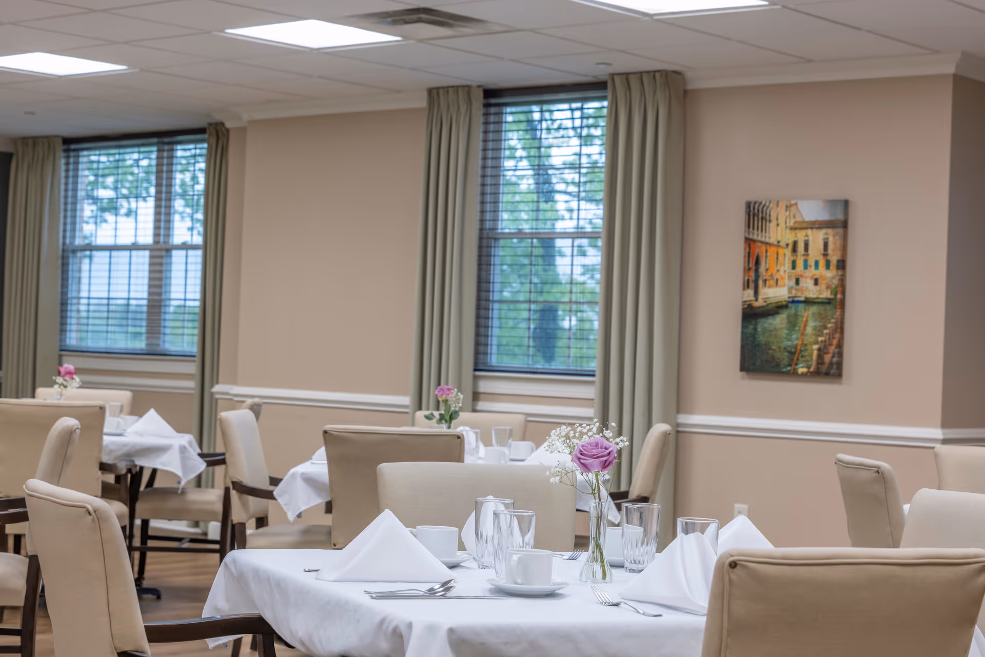 A dining room with several tables covered in white tablecloths, each set with white napkins, cups, glasses, and silverware. Beige upholstered chairs surround the tables. There are windows with blinds and beige curtains, and a framed picture of a canal scene on the wall.