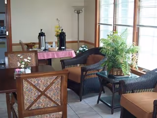 Sunlit common room with wicker and upholstered chairs, a potted fern on a side table, and a refreshment table with coffee urns near the windows.