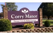 Outdoor view of a large purple sign for Corry Manor Therapy Care & Skilled Nursing, mounted between two brick pillars with greenery and trees in the background.
