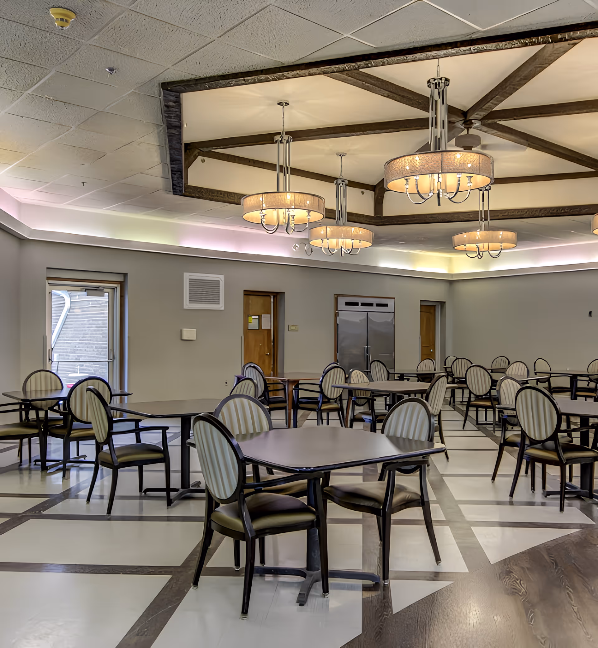 A spacious dining room with multiple tables and chairs arranged neatly. The chairs have striped upholstery and dark wooden frames. The ceiling features decorative wooden beams and several large, round chandeliers providing warm lighting. There are doors and a large stainless steel refrigerator along the far wall.