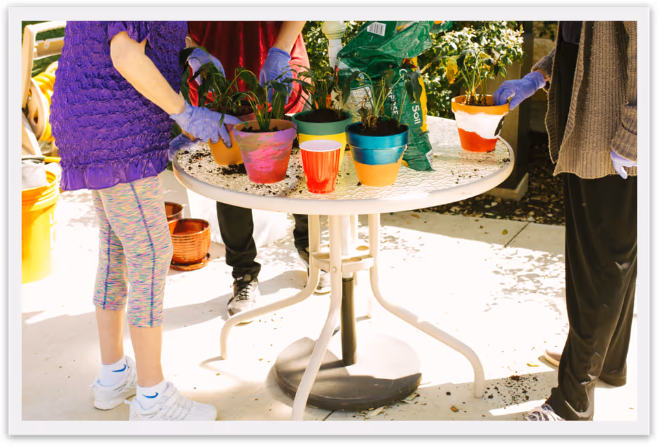 Three people wearing gloves stand around an outdoor table potting plants with colorful flowerpots and soil.