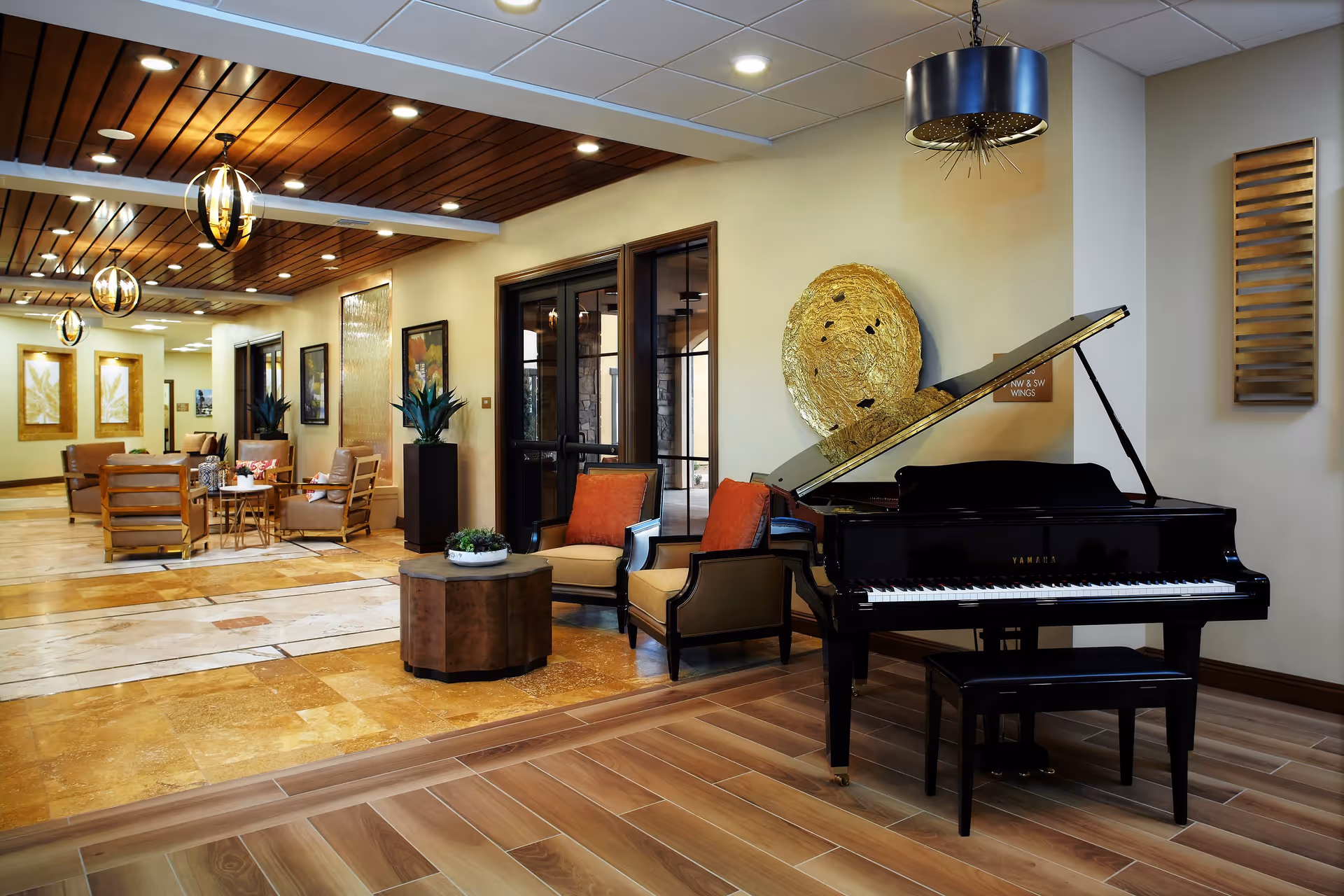 A spacious and well-lit interior common area featuring a black grand piano with a matching bench, two cushioned armchairs with orange pillows, a round wooden coffee table with a small plant, and additional seating with chairs and tables in the background. The ceiling has wooden paneling with modern hanging light fixtures, and the floor is a combination of wood and tile. Decorative wall art and plants add to the warm ambiance.