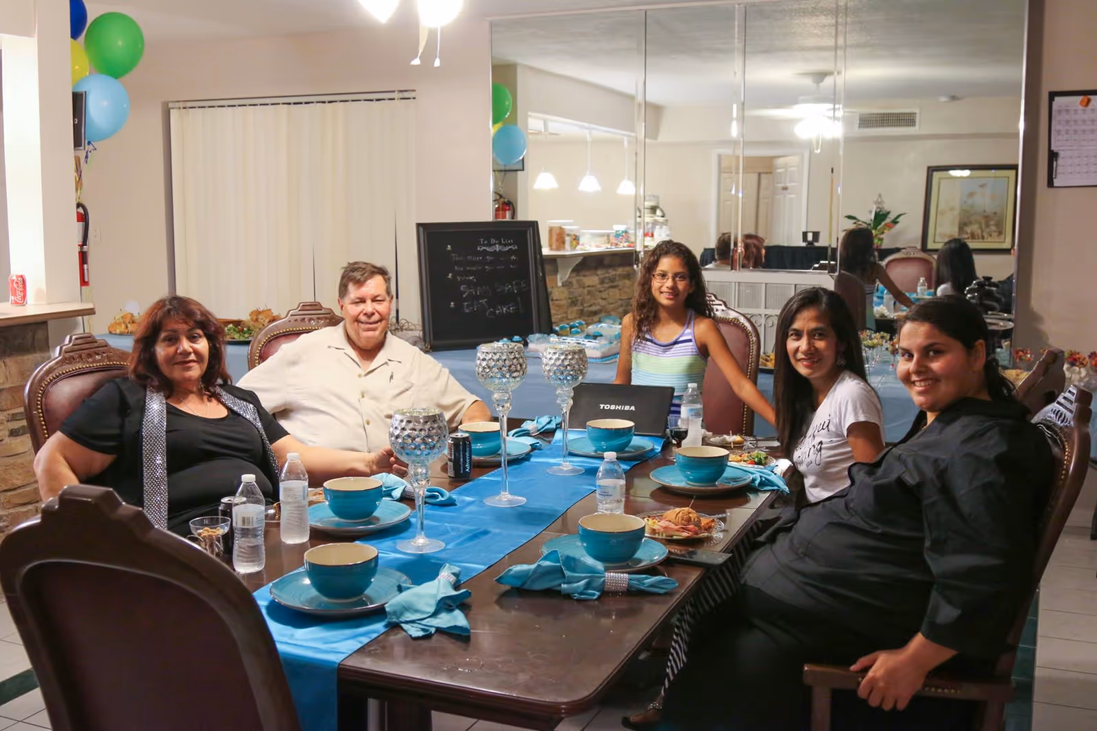 Five people sitting around a dining table set with blue table runner, turquoise bowls, plates, and napkins. The room has a large mirror on one wall, balloons in the corner, and a chalkboard in the background with some writing. The group appears to be enjoying a meal together in a cozy indoor setting.