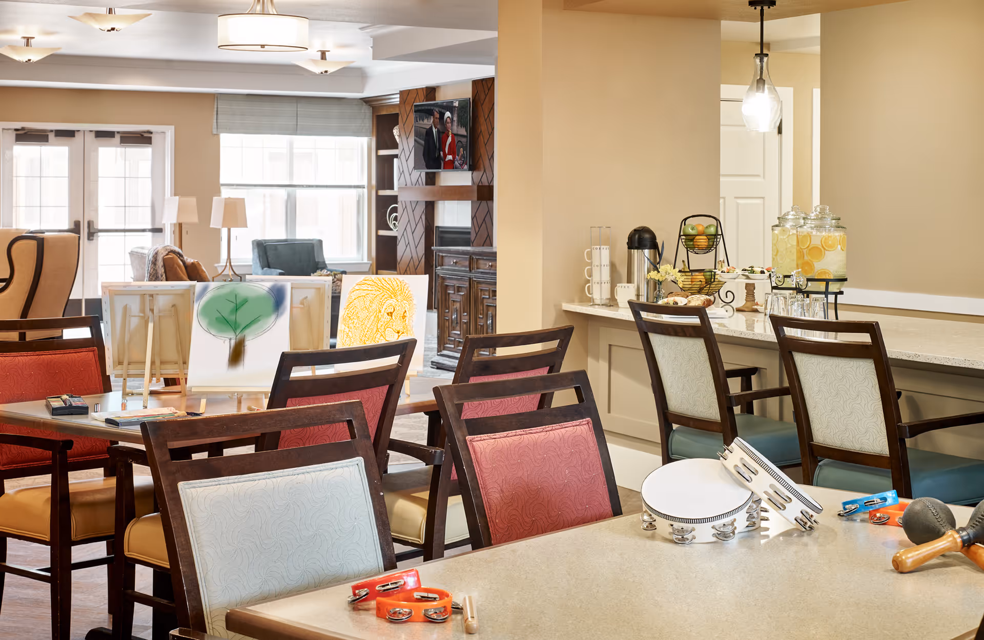 Bright communal activity and dining room with tables, chairs, easels, craft supplies, and a countertop beverage station.