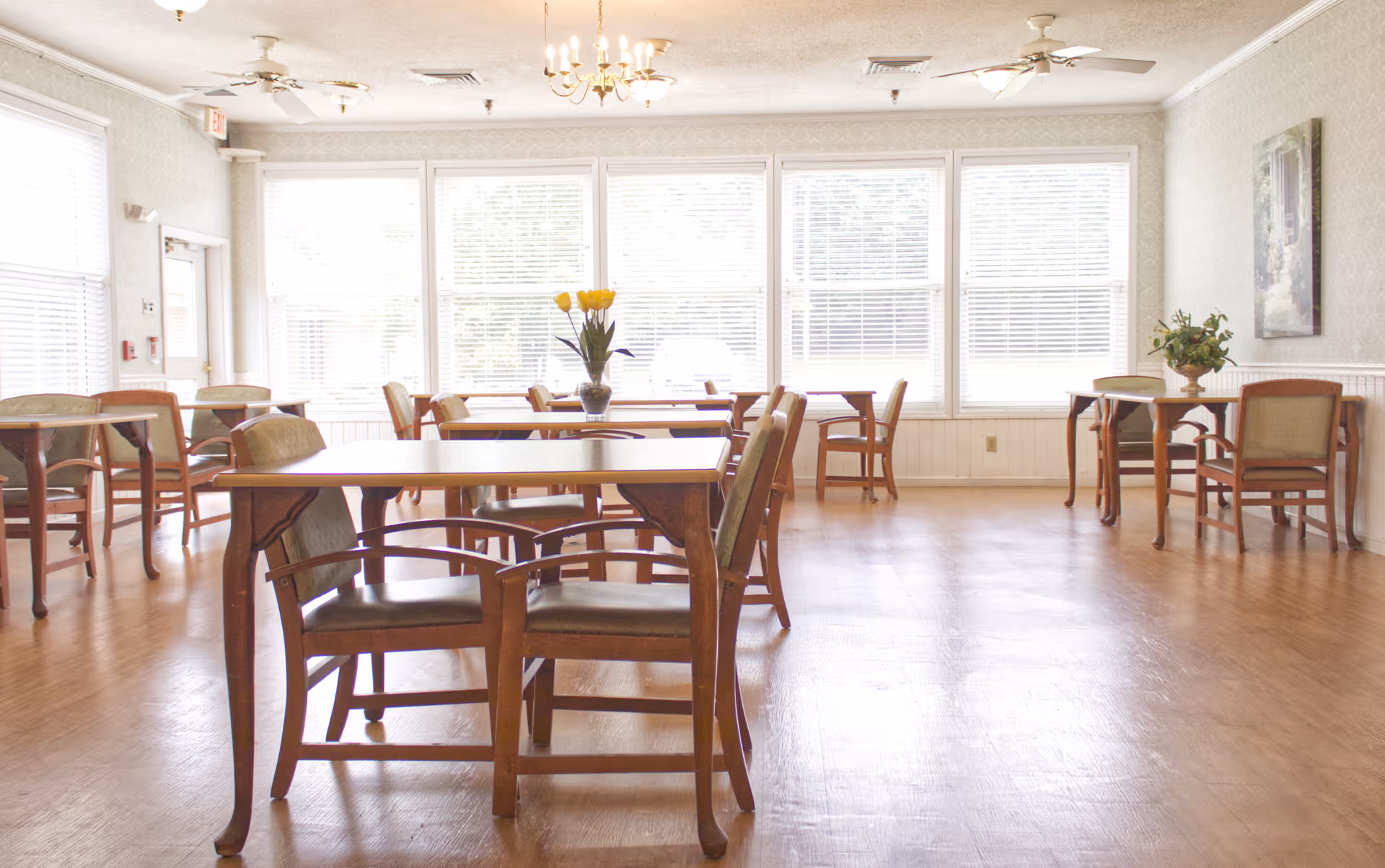 A bright dining room with multiple wooden tables and chairs arranged neatly. Large windows with white blinds allow natural light to fill the room. There are two vases with flowers placed on separate tables, and a chandelier and ceiling fans are visible on the ceiling.