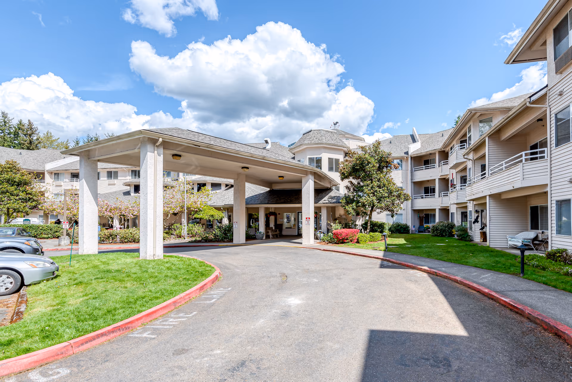 Exterior view of Solstice Senior Living at Renton showing the entrance driveway with a covered drop-off area, surrounded by a multi-story building with balconies, green lawns, trees, and a partly cloudy blue sky.