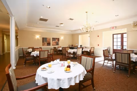 A bright dining room with several round tables covered with white tablecloths, each set with plates, cups, and glasses of orange juice. The room has beige walls, carpeted floors, and wooden chairs with striped cushions. A chandelier hangs from the ceiling, and there are two framed artworks on the far wall. Sunlight streams in through a glass door on the right side.