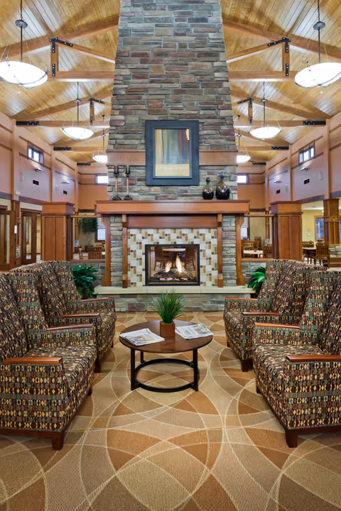 Cozy communal lounge with patterned armchairs arranged around a central coffee table facing a stone fireplace under a vaulted wooden ceiling.