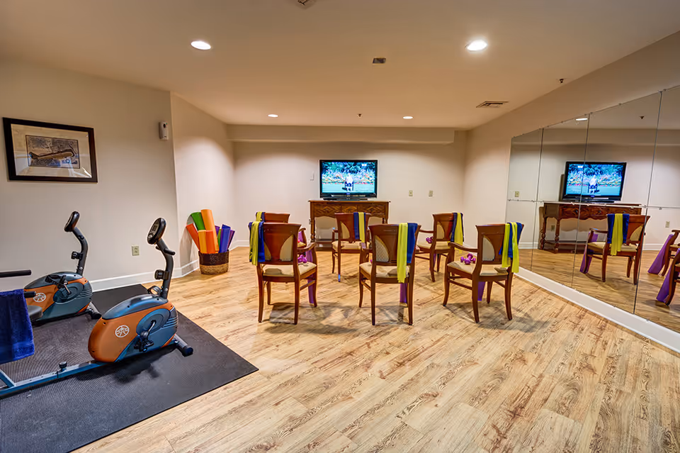 Exercise/activity room with two stationary bikes, chairs arranged facing a wall-mounted TV, mirrored wall and colorful exercise bands and mats.