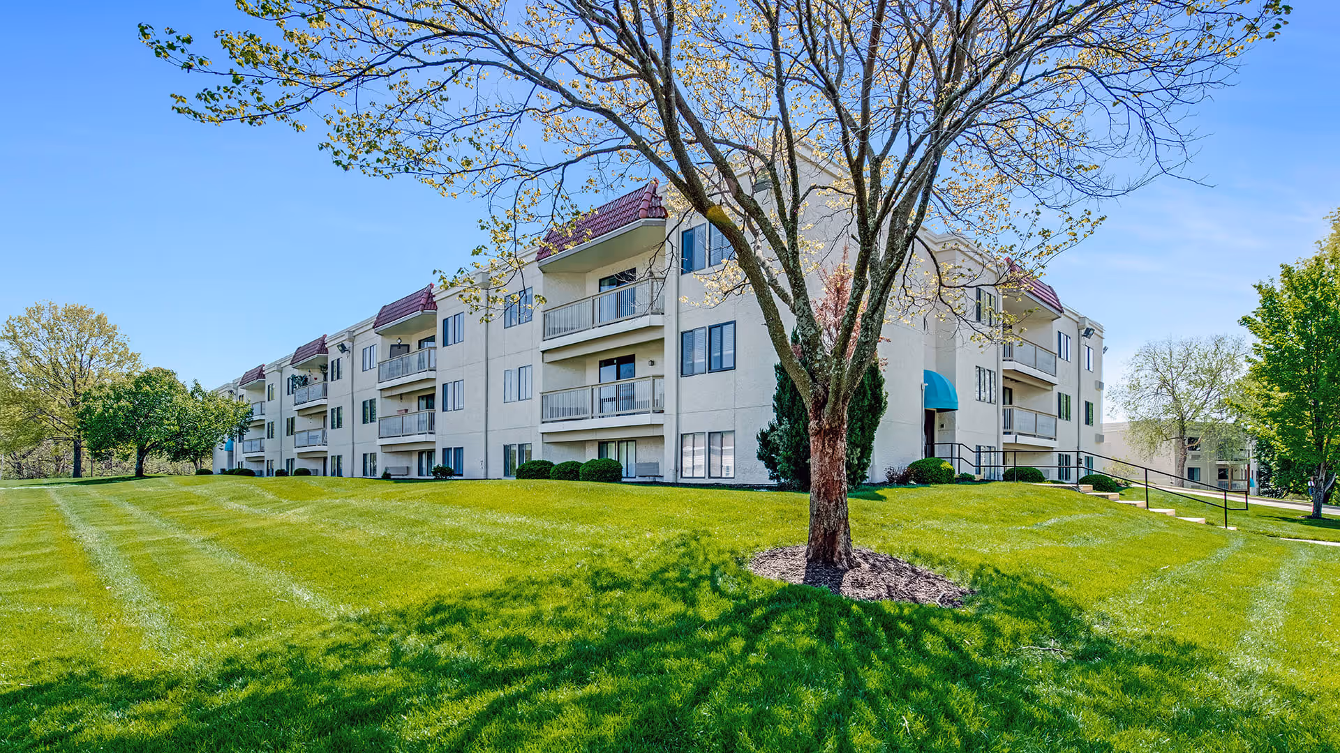 Exterior view of a multi-story residential building with balconies, surrounded by a well-maintained green lawn and trees under a clear blue sky.