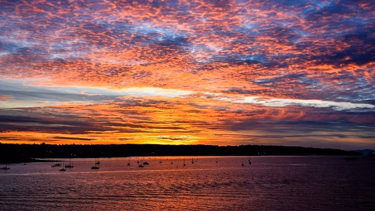 A vibrant sunset over a body of water with numerous small boats anchored near the shore and a dark silhouette of land in the background under a colorful sky filled with orange, pink, purple, and blue clouds.