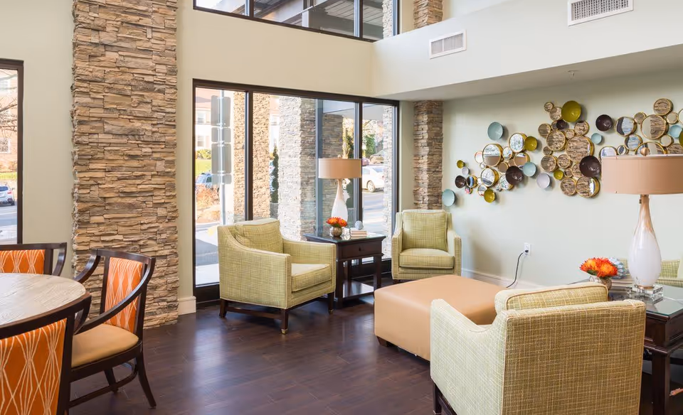 Bright communal seating area with green upholstered armchairs, a tan ottoman, a round dining table, decorative wall art and large floor-to-ceiling windows.