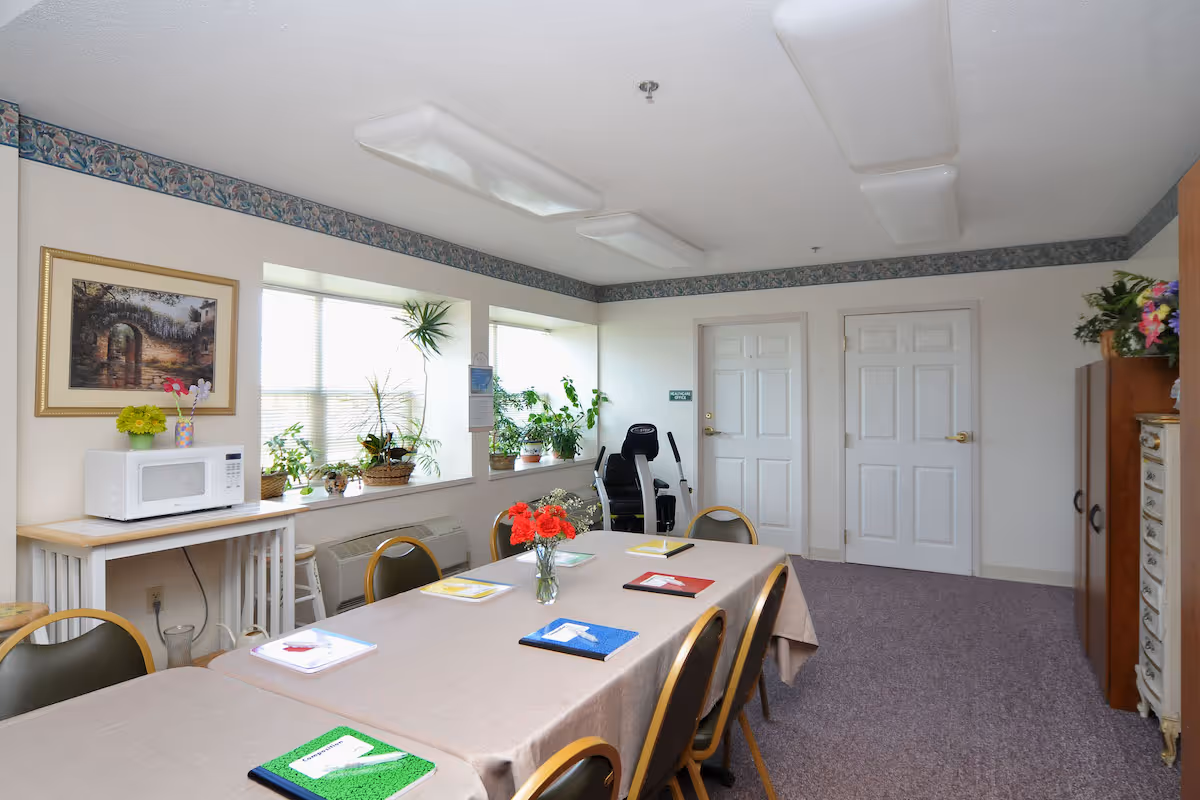 A well-lit room with a long table covered with a beige tablecloth and surrounded by chairs. On the table are colorful notebooks and a vase with red flowers. The room has large windows with several potted plants on the windowsill. There is a microwave on a small table to the left, and two closed white doors on the far wall. The room has a carpeted floor and floral wallpaper border near the ceiling.