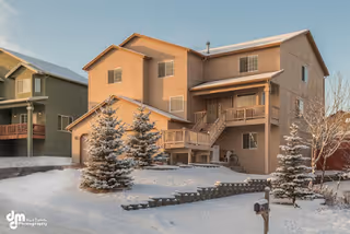 A two-story residential building covered in light brown siding with a snow-covered yard and evergreen trees in front. The scene is set in winter with snow on the ground and trees, and the sky is clear.