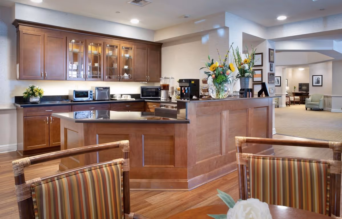 A bright communal kitchen and seating area with a wooden island, cabinets, appliances, floral arrangements, and lounge seating visible in the background.