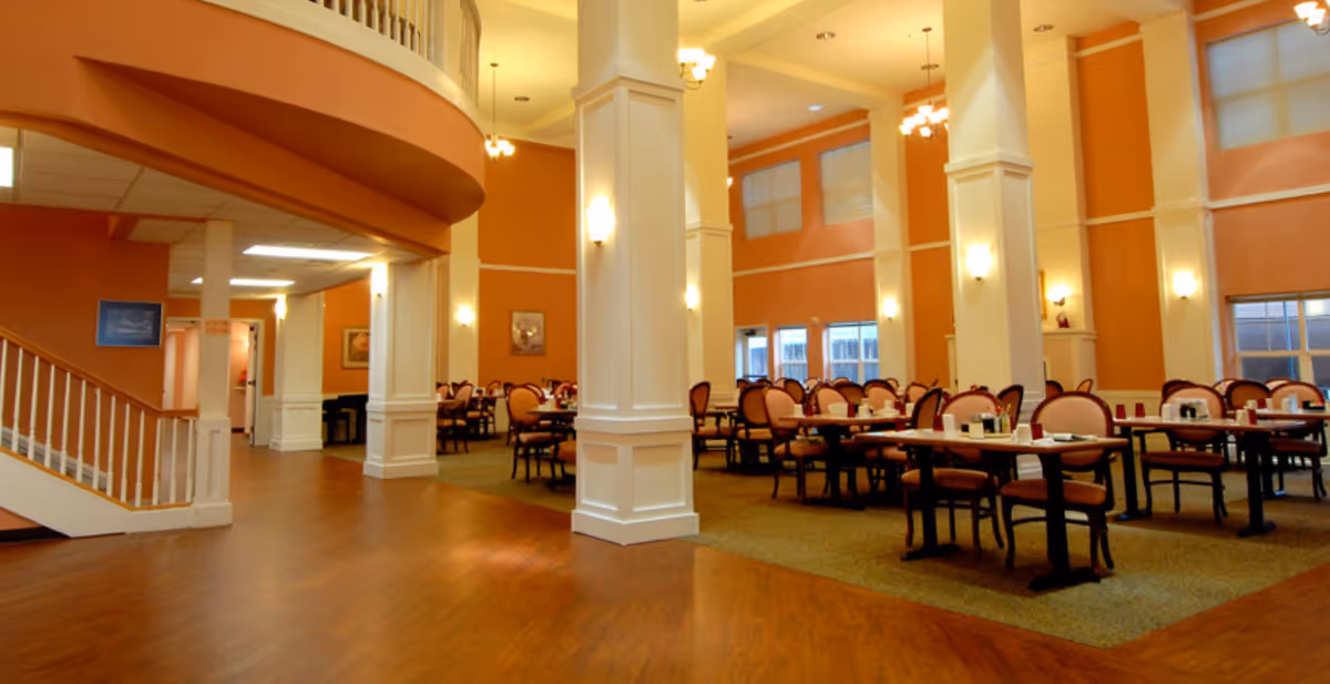 Spacious dining room in The Bridge Retirement & Assisted Living with multiple tables and chairs arranged neatly. The room features high ceilings, large white columns, warm orange walls, and several windows allowing natural light to enter. There is a staircase on the left side and soft lighting fixtures on the walls and ceiling.